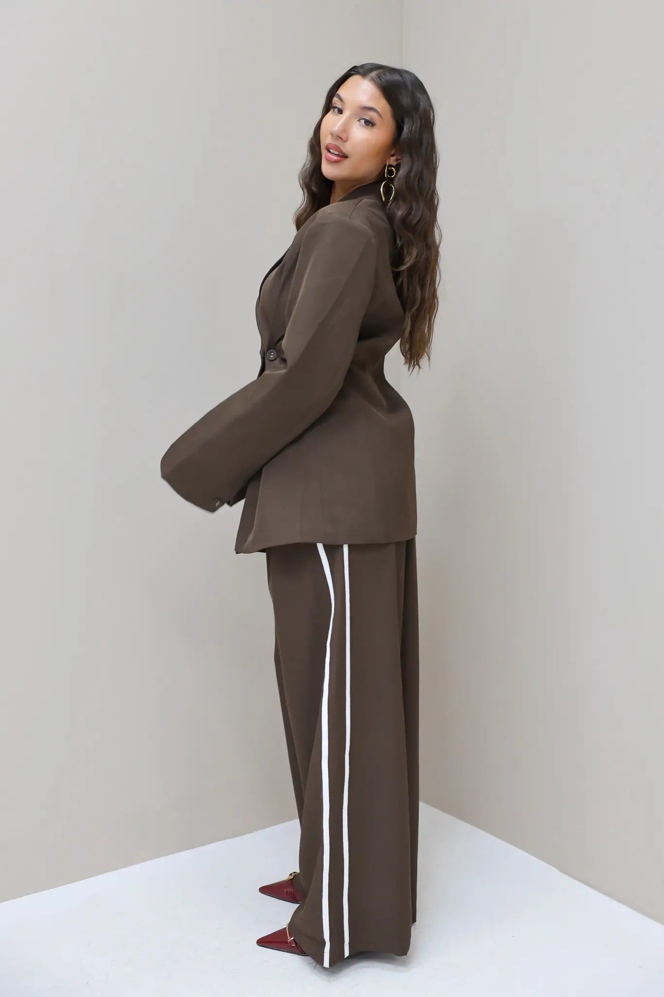 Model kijkt schuin over schouder in donkerbruine blazer met pantalon met witte bies