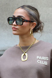 close-up van model met donkergroene zonnebril, gouden ketting en trui met Paris Club logo
