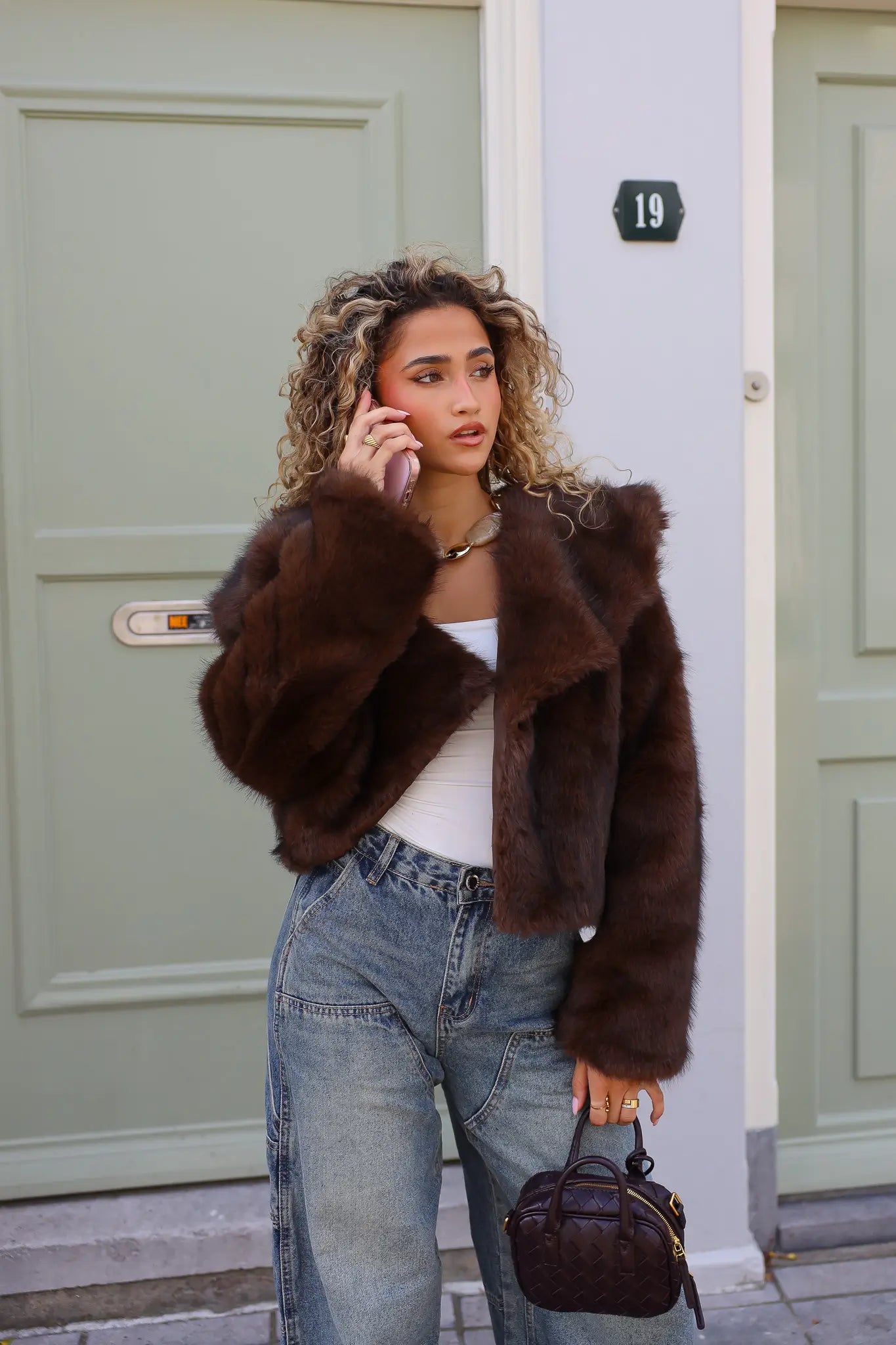 model draagt bruin faux fur jasje buiten voor een groene deur