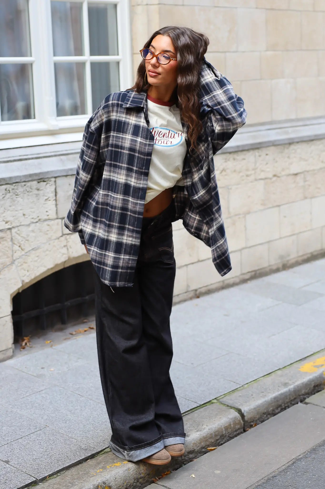 Streetstyle outfit met een open donkerblauwe geruite flanellen blouse, een crème shirt met print en zwarte wide leg jeans.