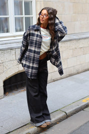 Streetstyle outfit met een open donkerblauwe geruite flanellen blouse, een crème shirt met print en zwarte wide leg jeans.