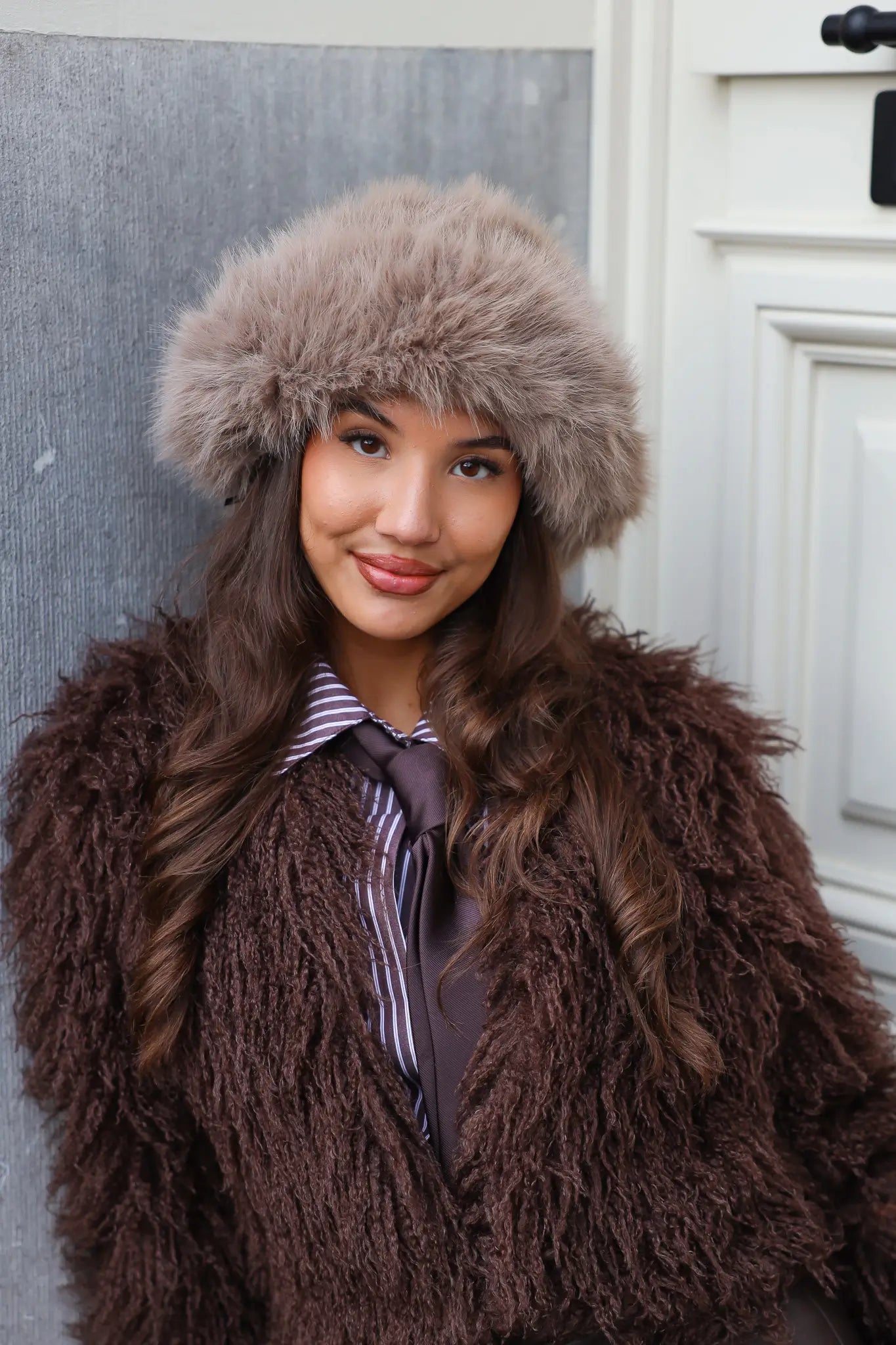 Model leunt tegen muur met fluffy muts en bruine jas