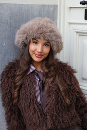 Model leunt tegen muur met fluffy muts en bruine jas