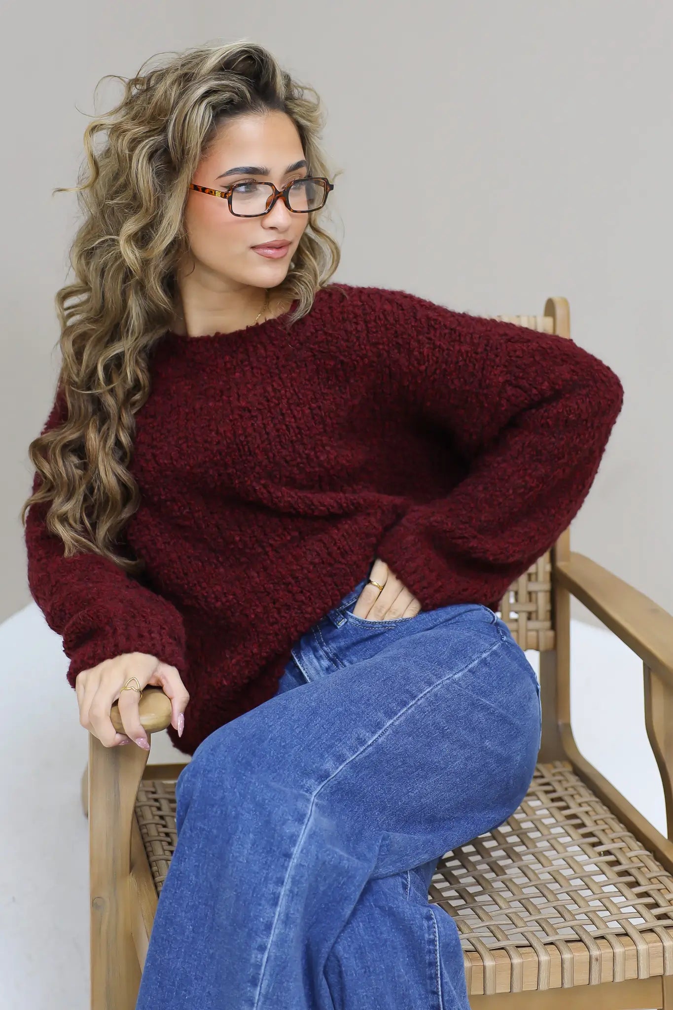 Model zit op een houten stoel met bordeaux trui en spijkerbroek
