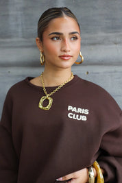 model draagt gouden ketting met hanger bij bruine trui, kijkt opzij