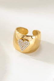 close-up van gouden ring met hartvormig design vol kleine steentjes op witte ondergrond