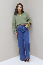 Model staat recht in groene fluffy trui met blauwe jeans en hakken