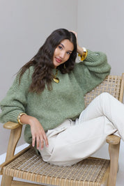 Model zit half liggend op stoel in groene fluffy trui met witte broek