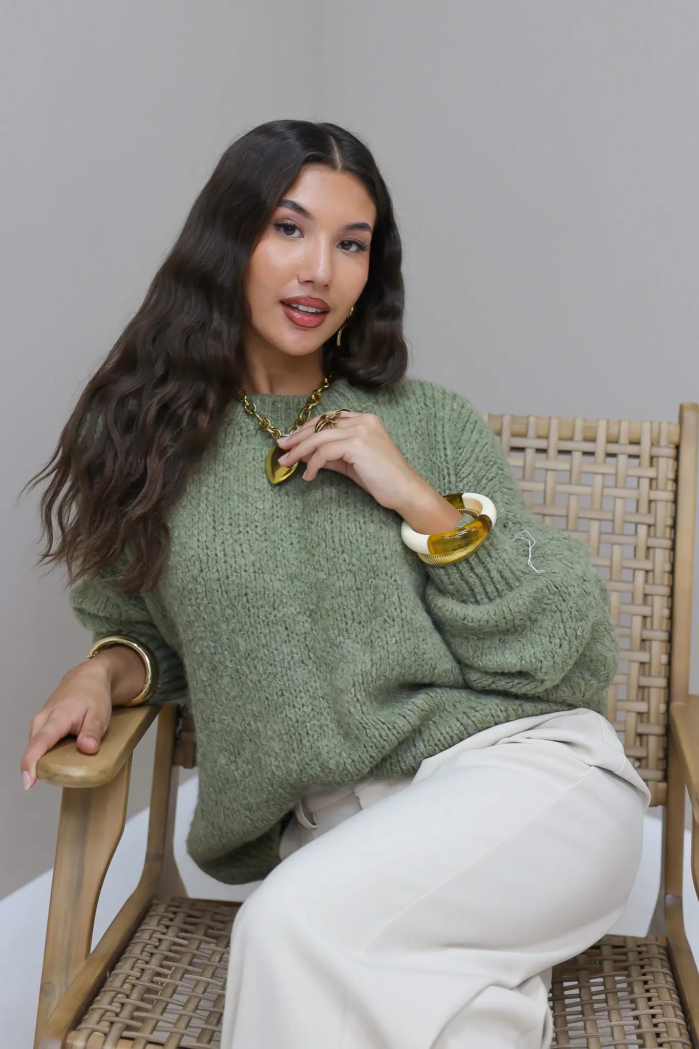 Model zit op stoel in groene fluffy trui met witte broek en gouden sieraden