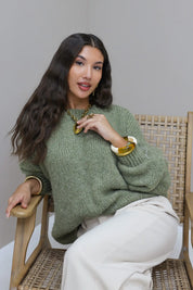 Model zit op stoel in groene fluffy trui met witte broek en gouden sieraden