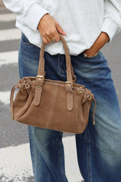 Taupe handtas met suèdelook, dubbele hengsels, gespen en trekkoorddetails, gecombineerd met een witte sweater en blauwe jeans.