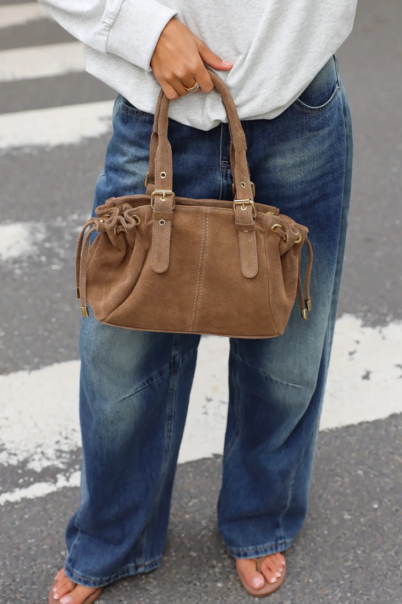 Taupe handtas met dubbele hengsels en goudkleurige details, gedragen bij een witte sweater en blauwe jeans.