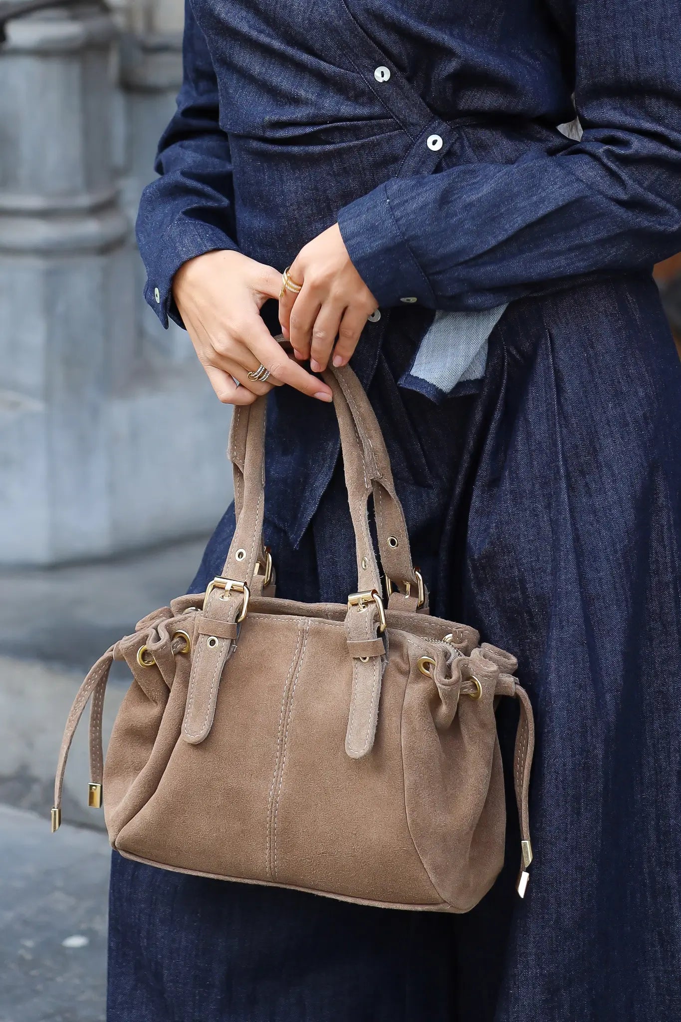 Detailfoto van een taupe handtas met suèdelook, goudkleurige gespen en dubbele hengsels, gedragen bij een donkerblauwe outfit.