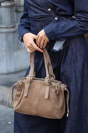 Detailfoto van een taupe handtas met suèdelook, goudkleurige gespen en dubbele hengsels, gedragen bij een donkerblauwe outfit.