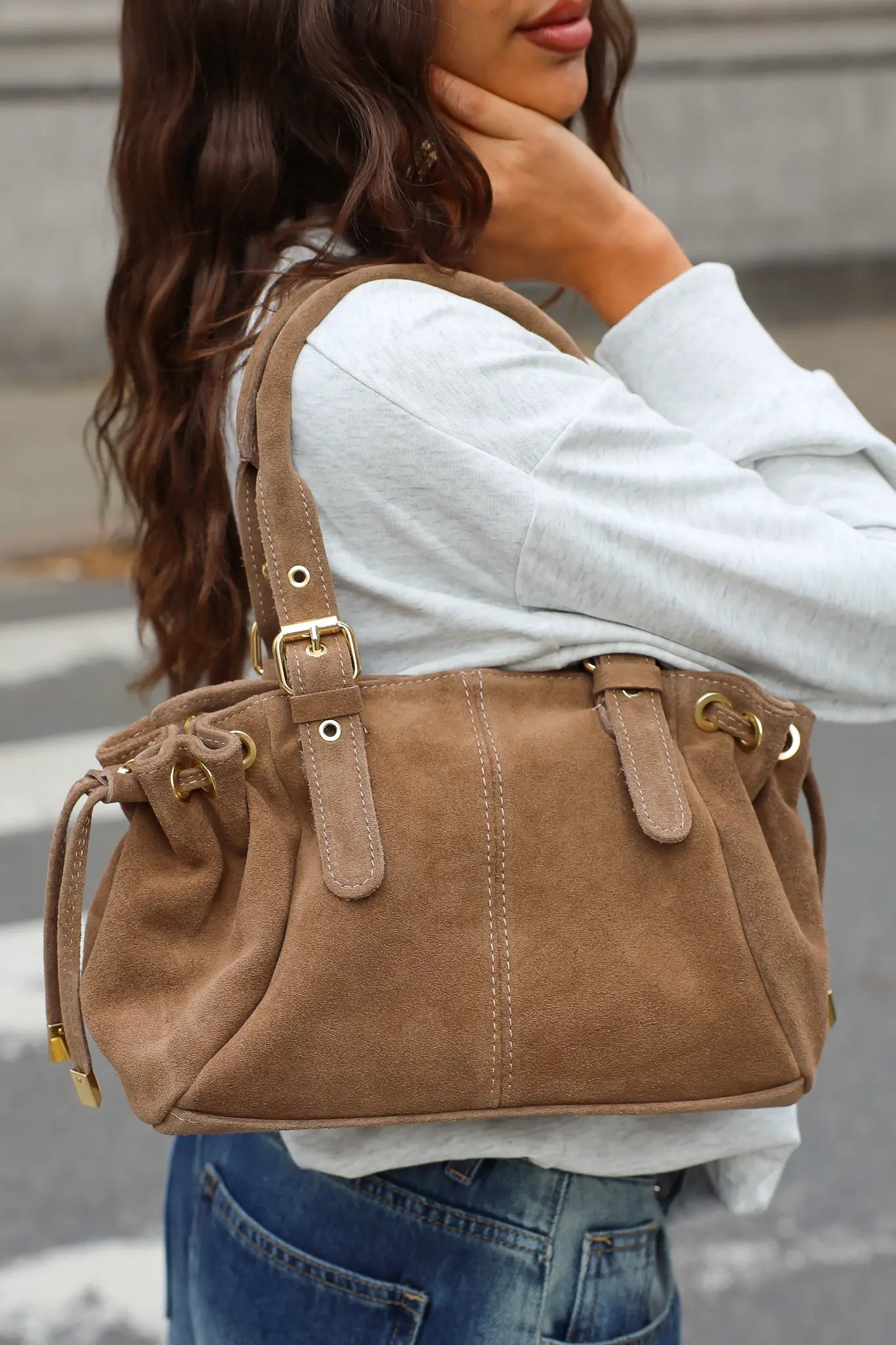 Close-up van een taupe schoudertas met suèdelook, dubbele hengsels, gespen en trekkoorddetails.