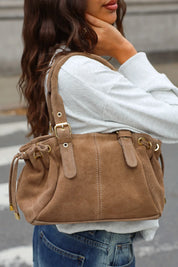 Close-up van een taupe schoudertas met suèdelook, dubbele hengsels, gespen en trekkoorddetails.