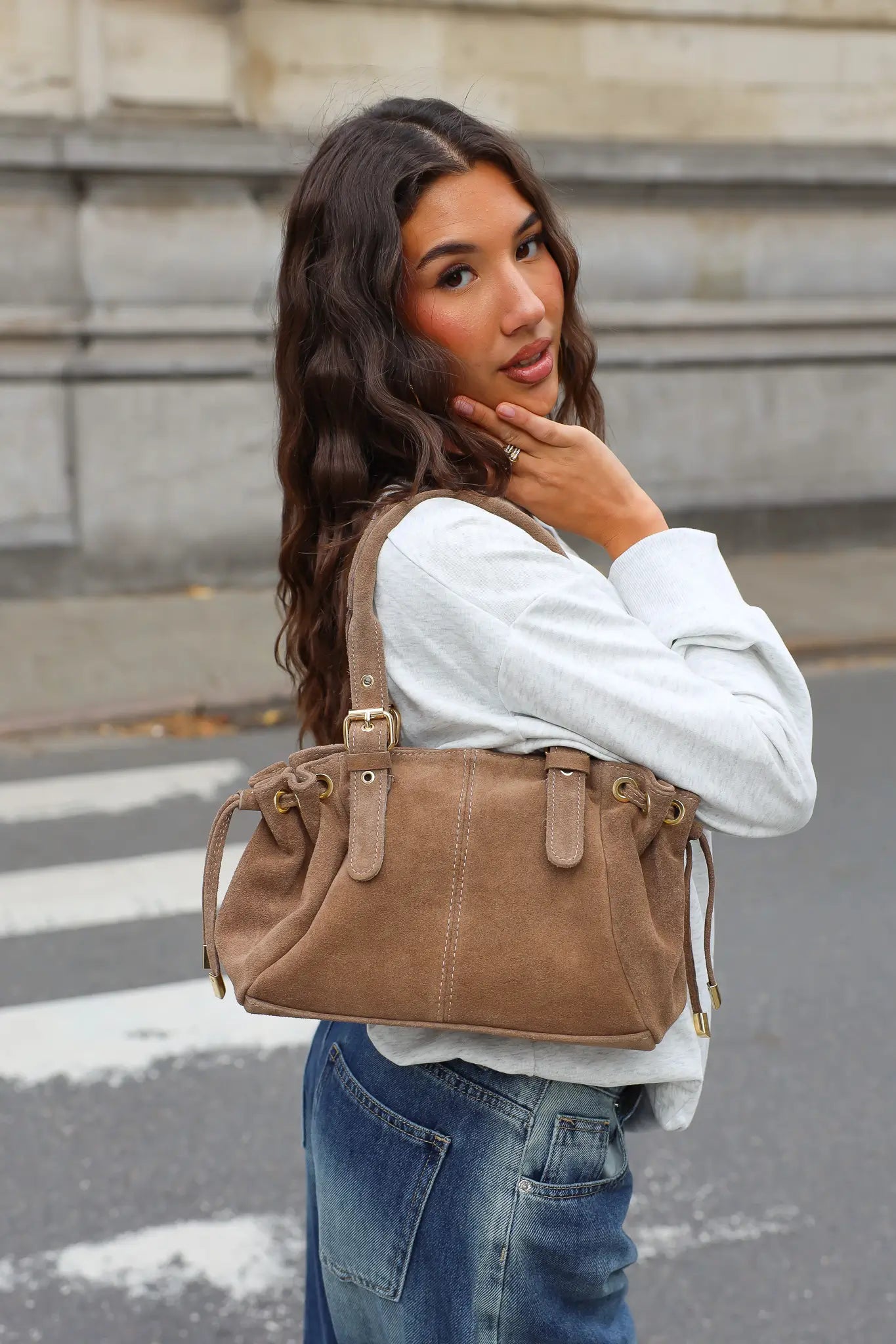 Taupe handtas met suèdelook, gedragen over de schouder bij een witte sweater en blauwe jeans.