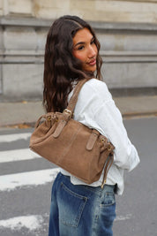 Taupe handtas met suèdelook over de schouder gedragen bij een witte sweater en blauwe jeans, gefotografeerd vanaf de zijkant.