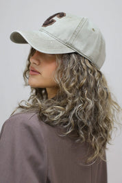 vrouw met krullen draagt beige honkbalpet met bruine letter c en kijkt over schouder