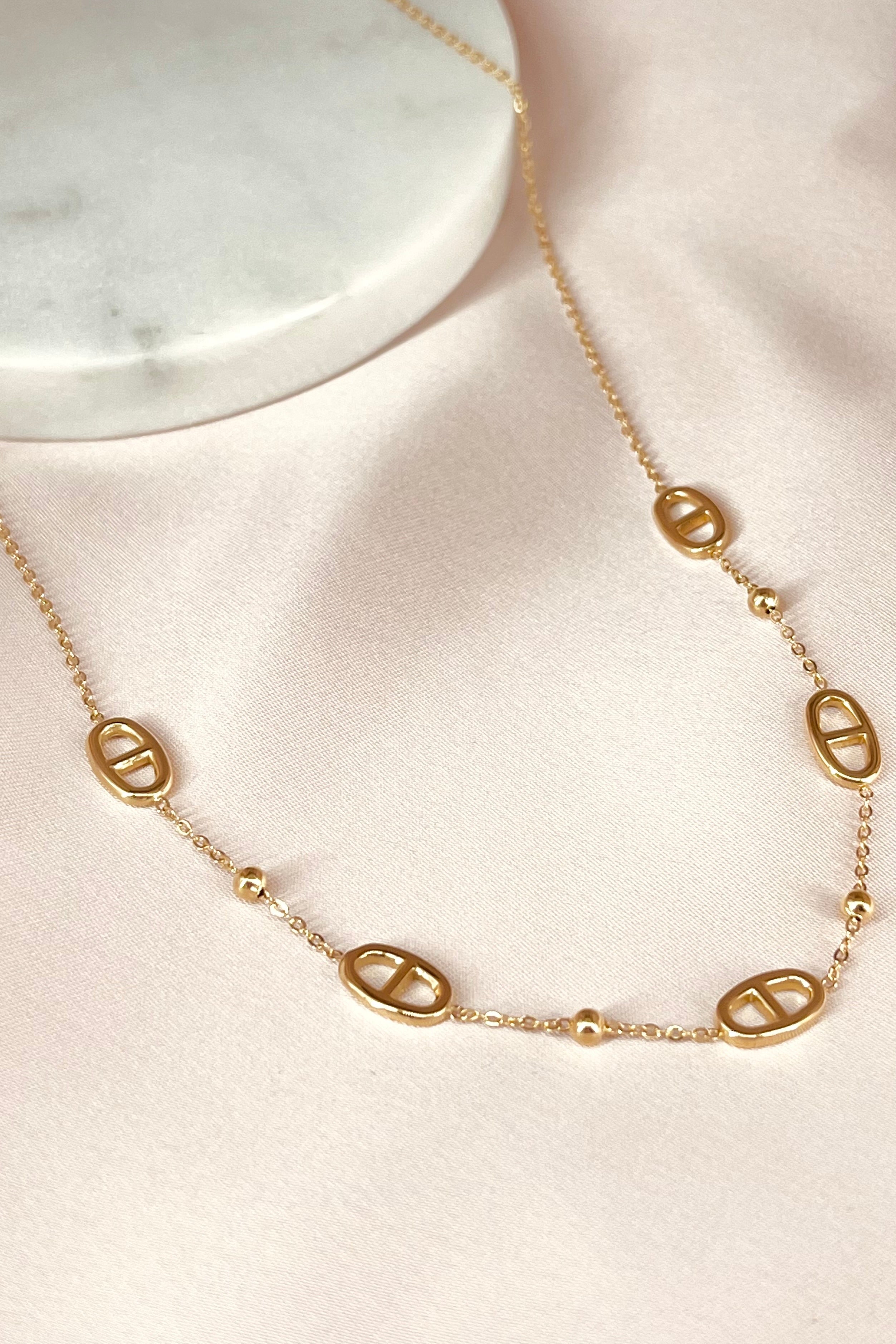 Gouden halsketting met ovalen schakels en kleine pareldetails op zacht roze achtergrond