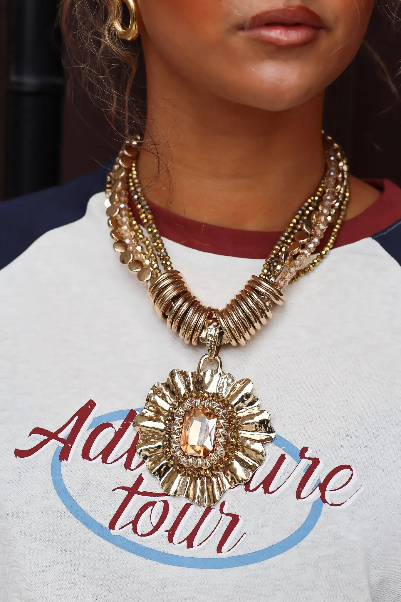 Close-up van een gouden layered statement ketting met grote ronde hanger gedragen bij een shirt met print