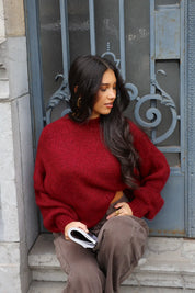 Model poseert zittend bij hek in de Liva trui bordeaux met bruine broek