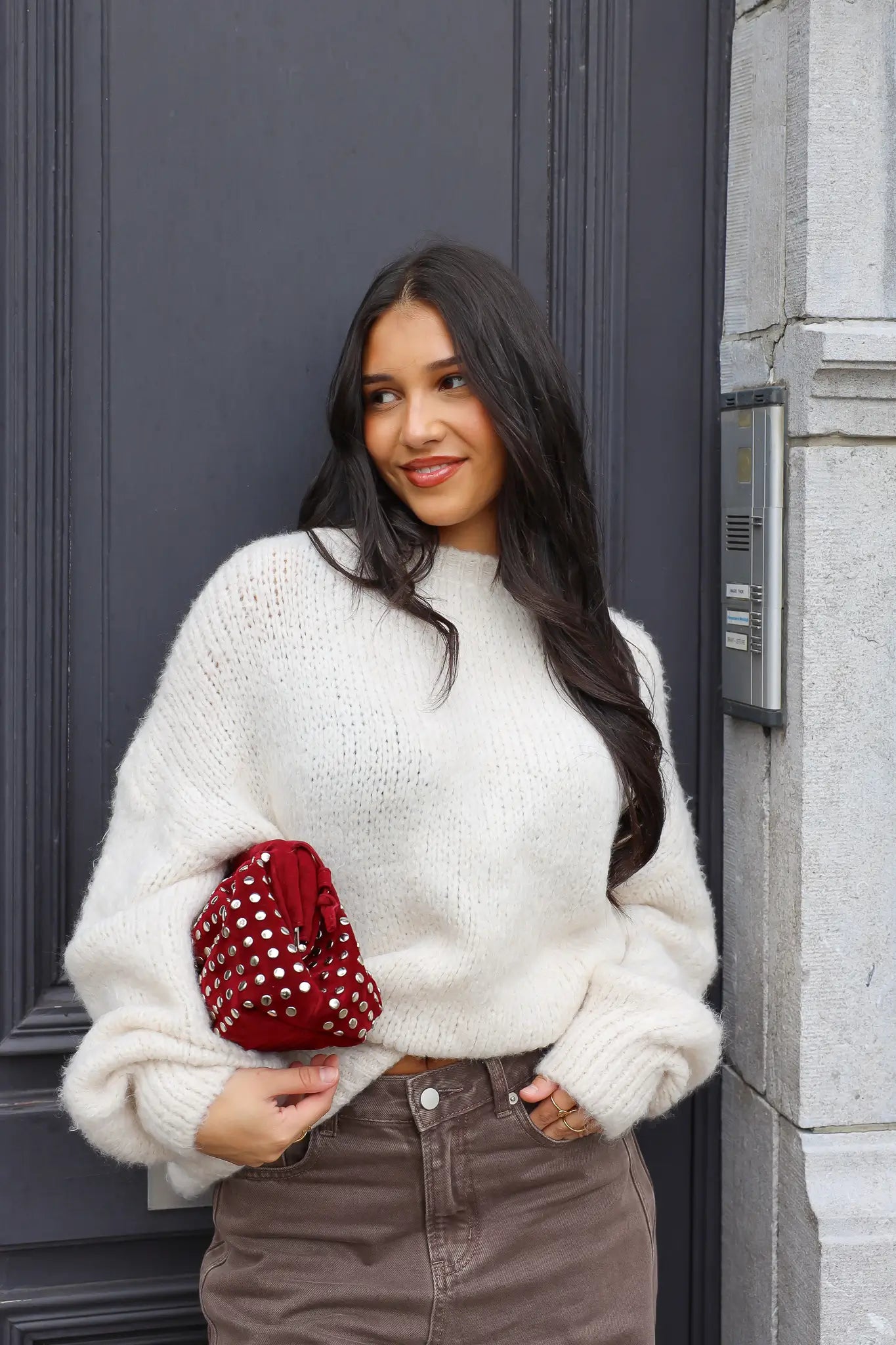 Model draagt de Liva trui wit met bruine broek en rode tas bij zwarte deur