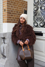 Model met beige muts en bruine jas buiten voor een deur