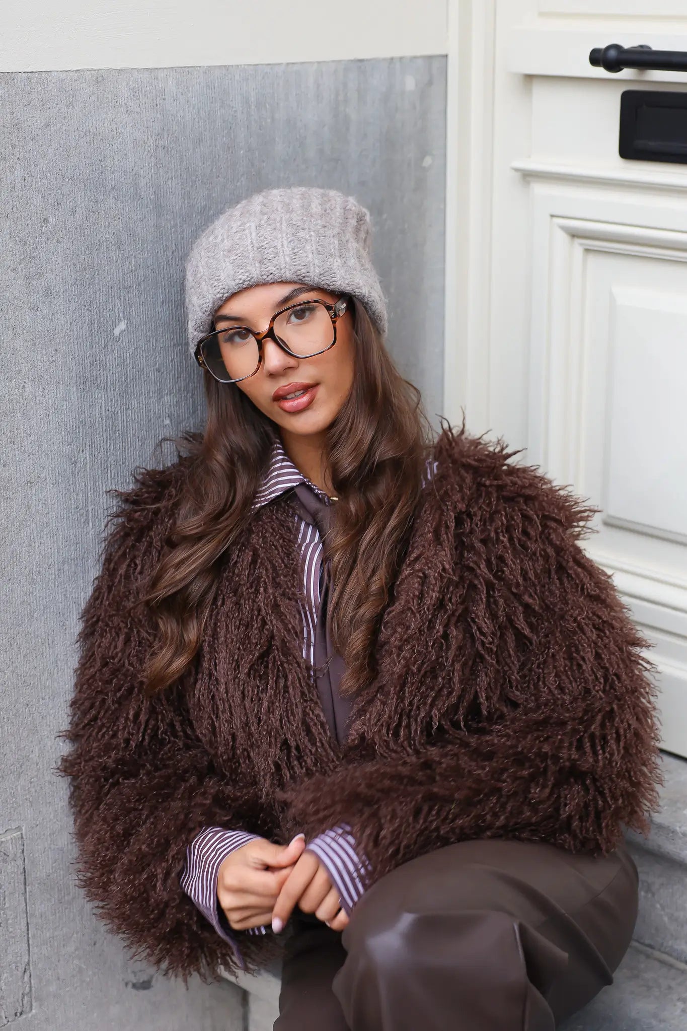 Model met taupe muts en bruine jas leunend tegen muur