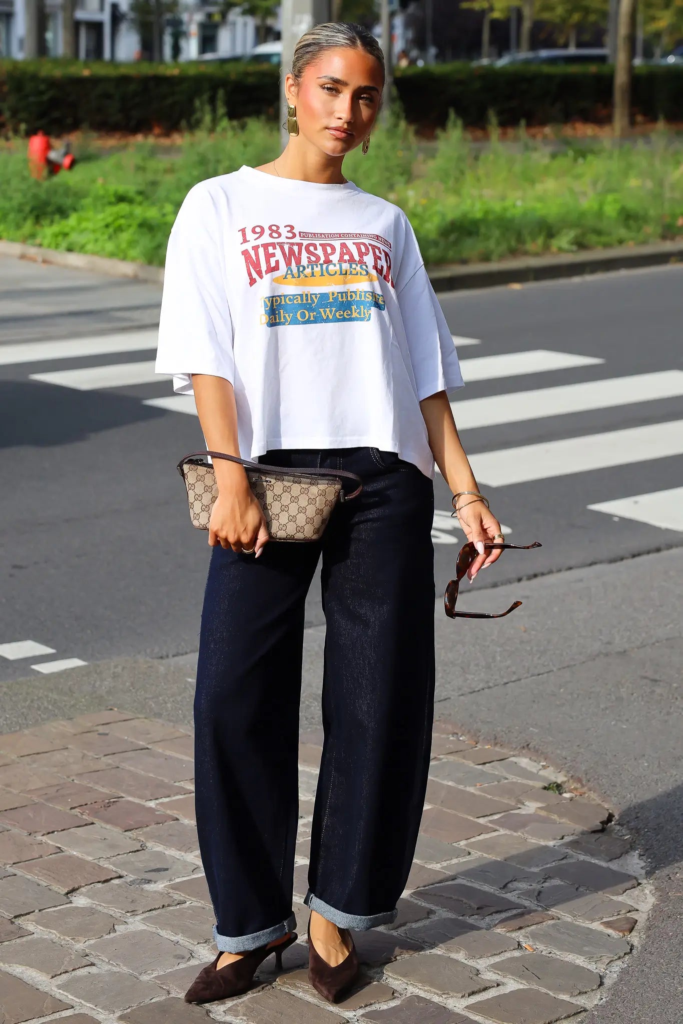 Wit newspaper T-shirt met kleurrijke opdruk gecombineerd met een zwarte wide leg jeans en beige tas tijdens een streetstyle fotoshoot