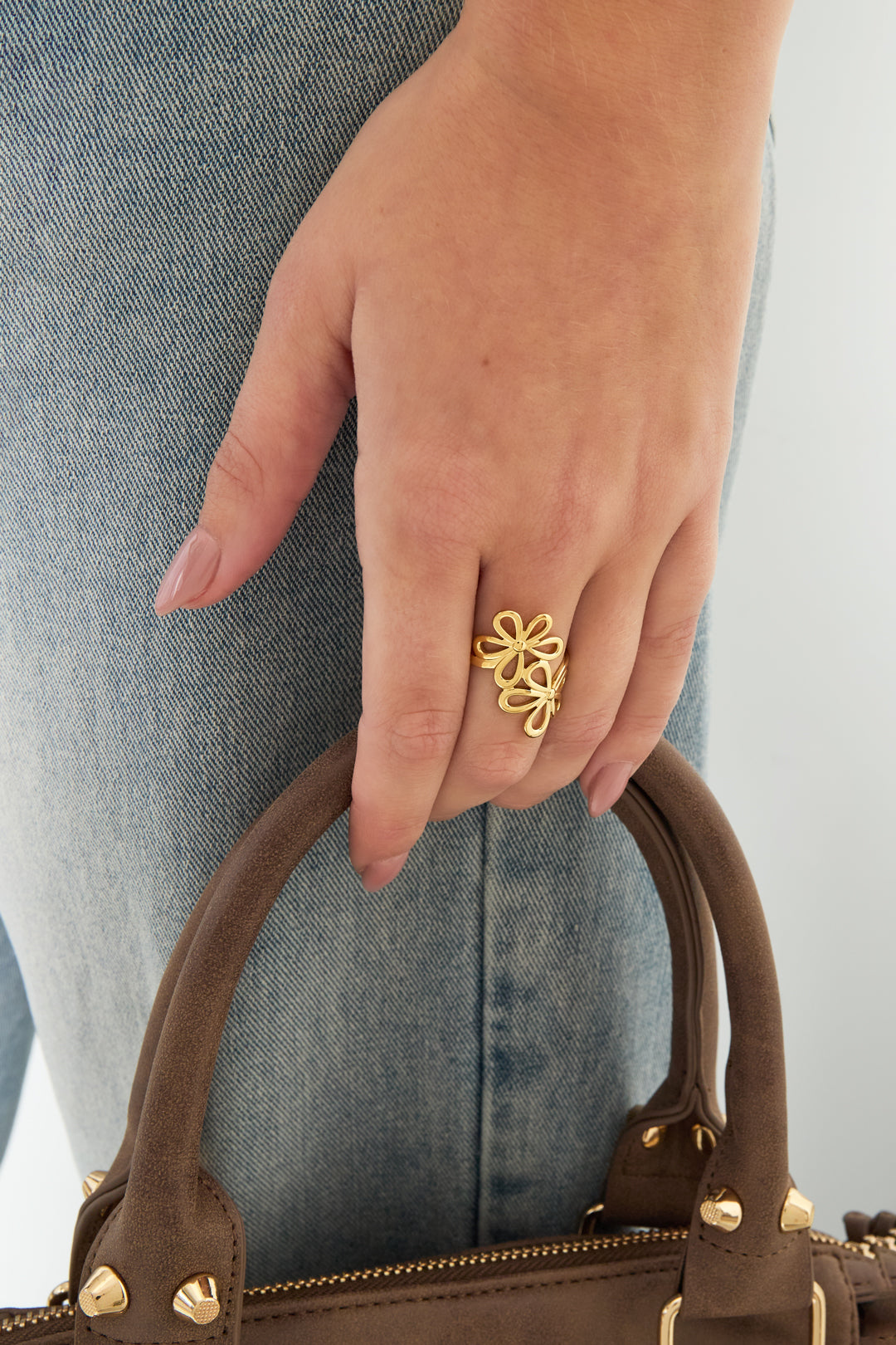 Hand met gouden bloemen ring bij spijkerbroek en bruine tas