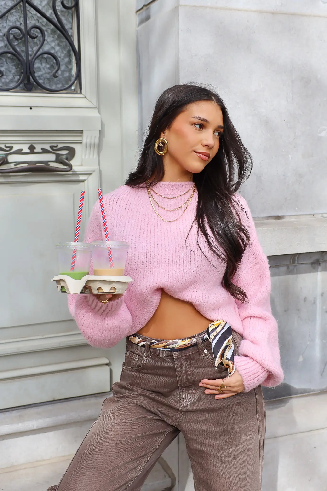 Model staat buiten in roze fluffy trui met bruine broek en houdt bekers vast