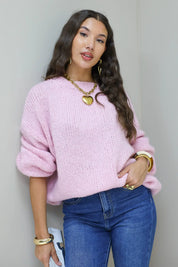 Model staat recht in roze fluffy trui met blauwe jeans en gouden sieraden