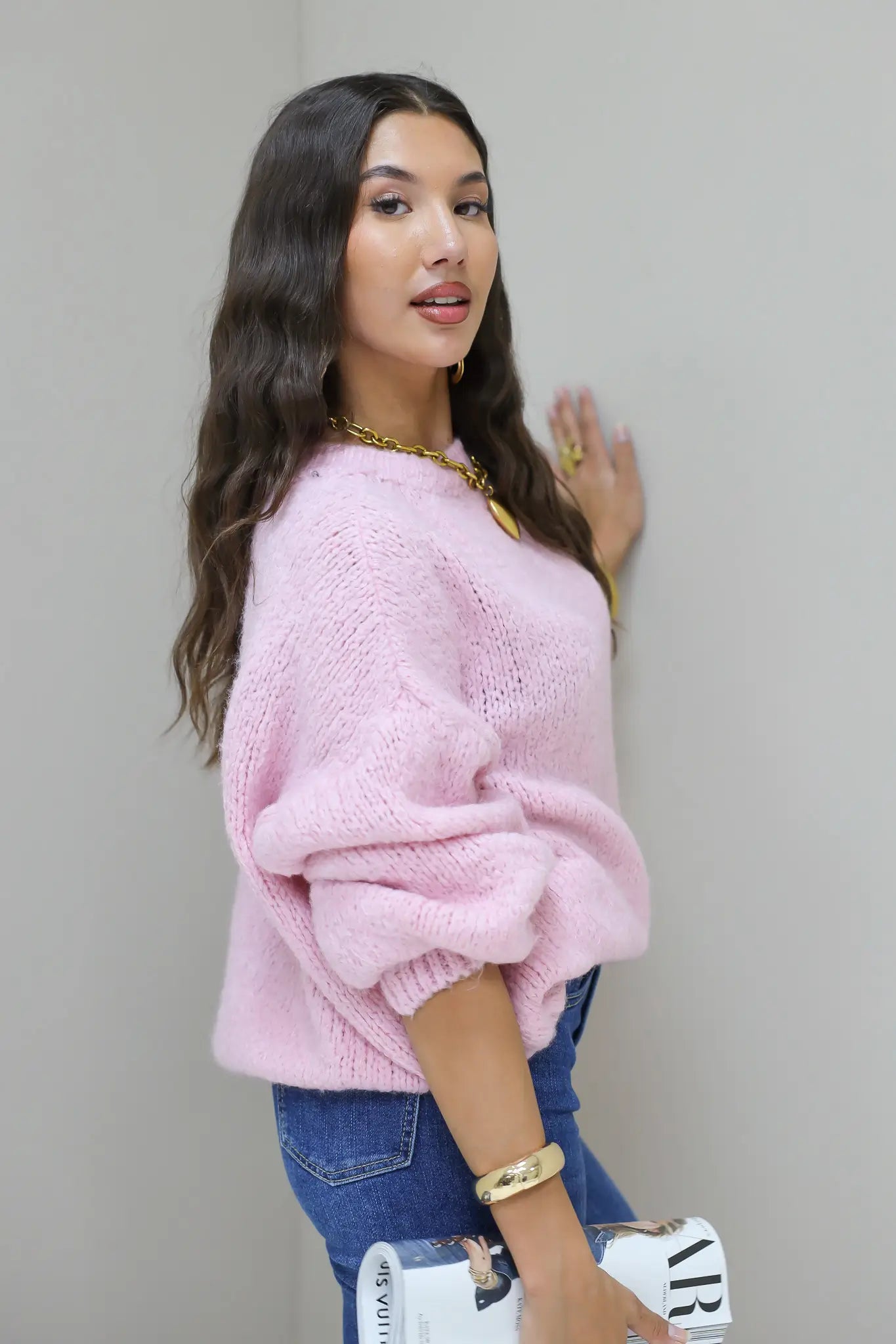 Model draait licht opzij in roze fluffy trui met blauwe jeans en witte tas in hand