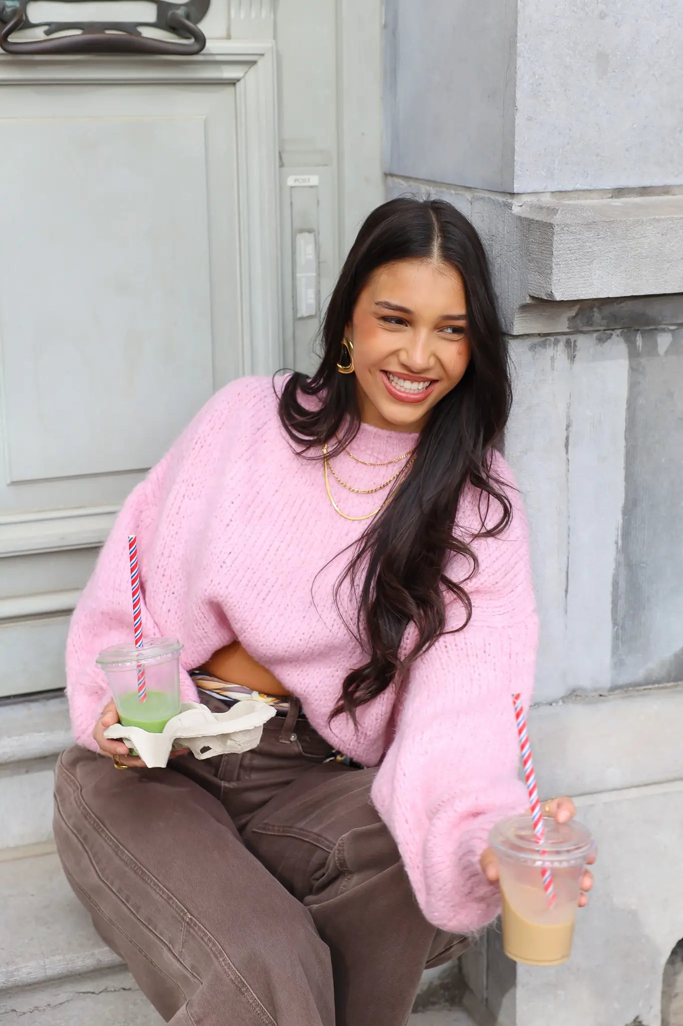 Model lacht op trap in roze fluffy trui met bruine broek en drankje