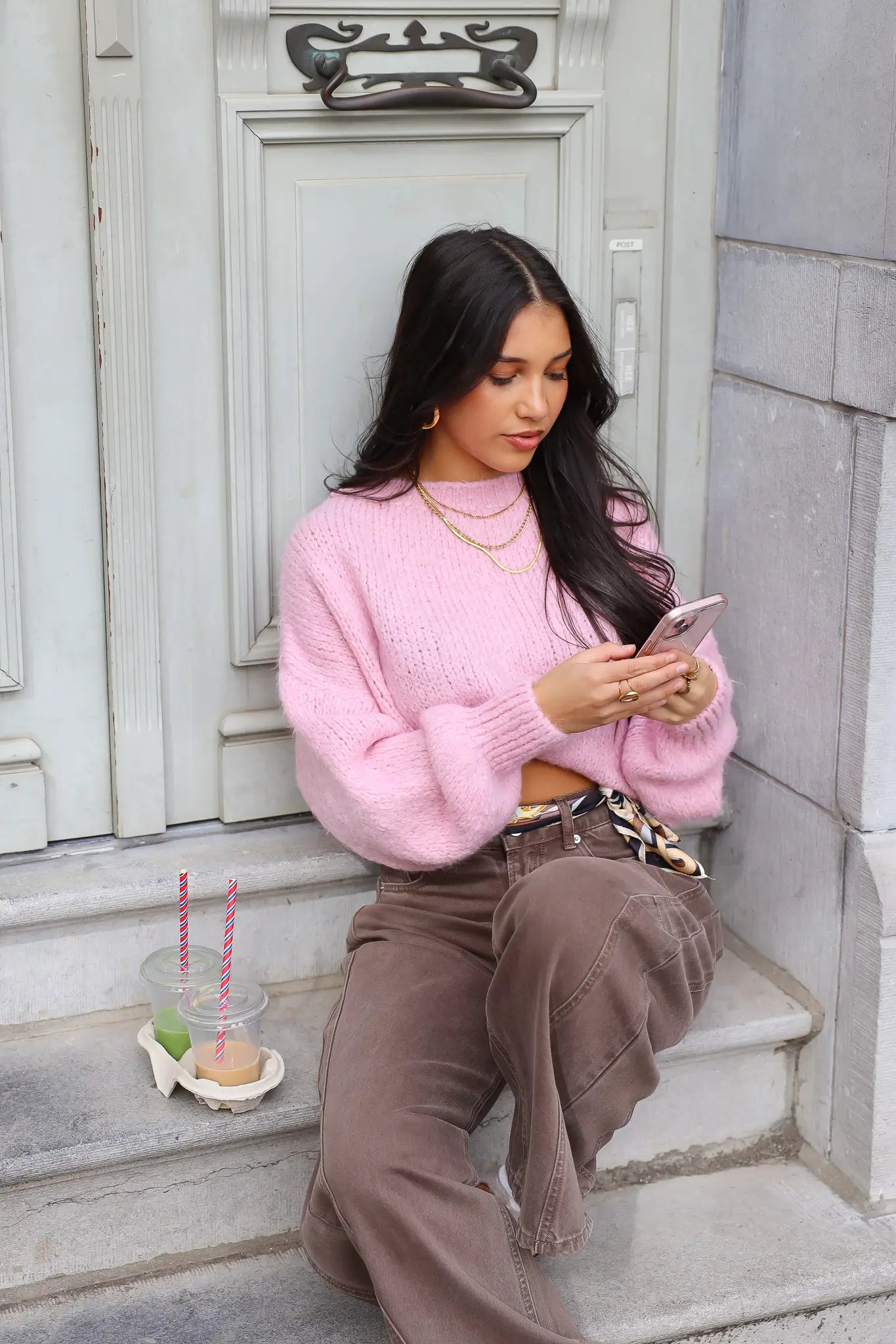 Model zit op traptreden in roze fluffy trui met bruine broek en bekers in hand