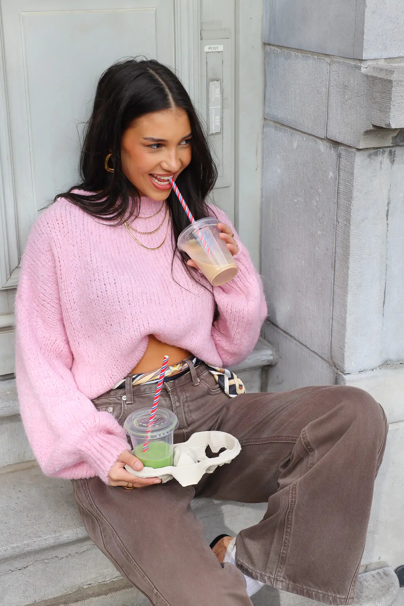 Model zit op trap met roze fluffy trui, bruine broek en kijkt opzij