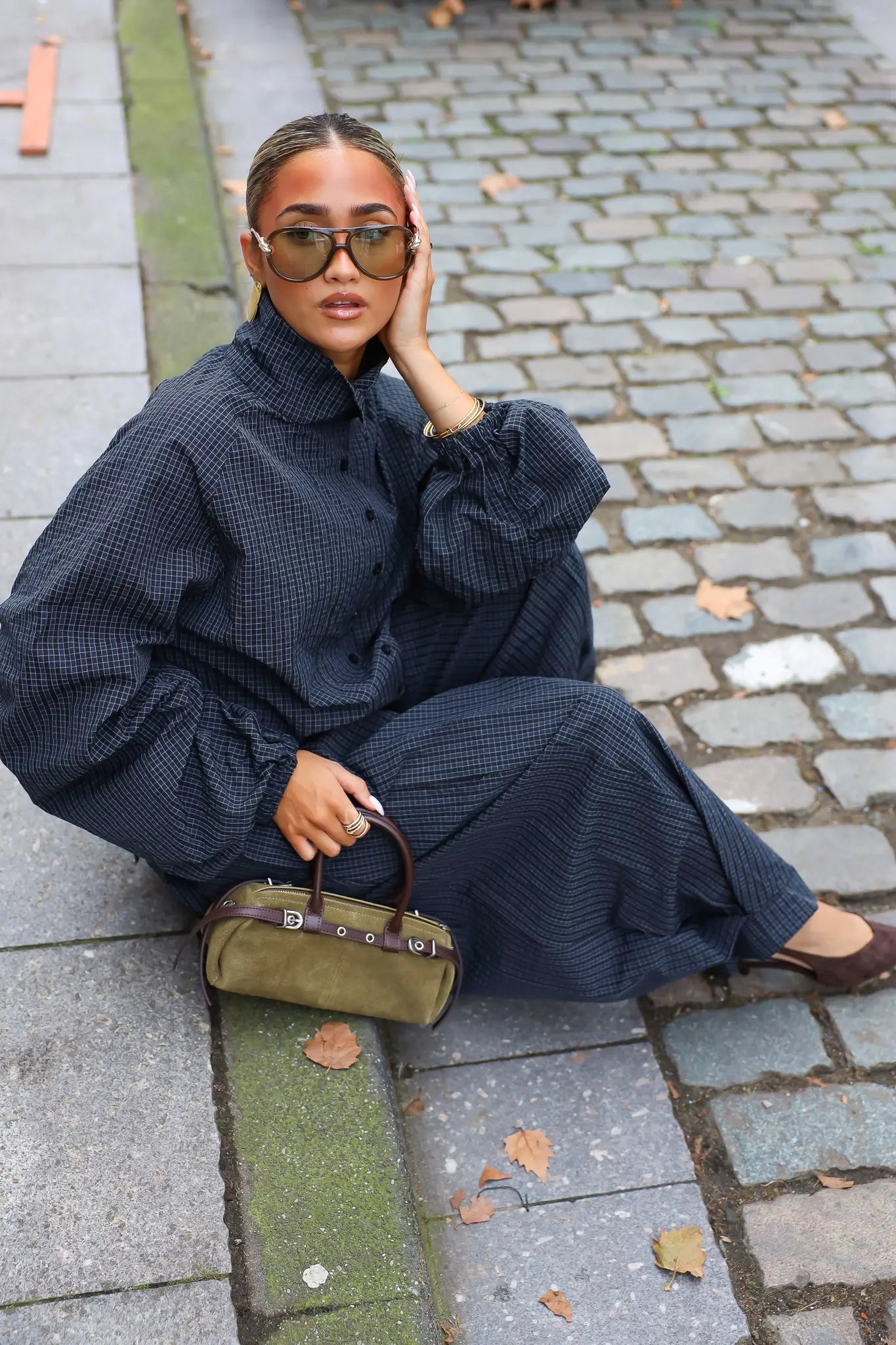 Khaki suède handtas met bruine details gecombineerd met een donkerblauwe oversized outfit en bruine schoenen tijdens een streetstyle fotoshoot