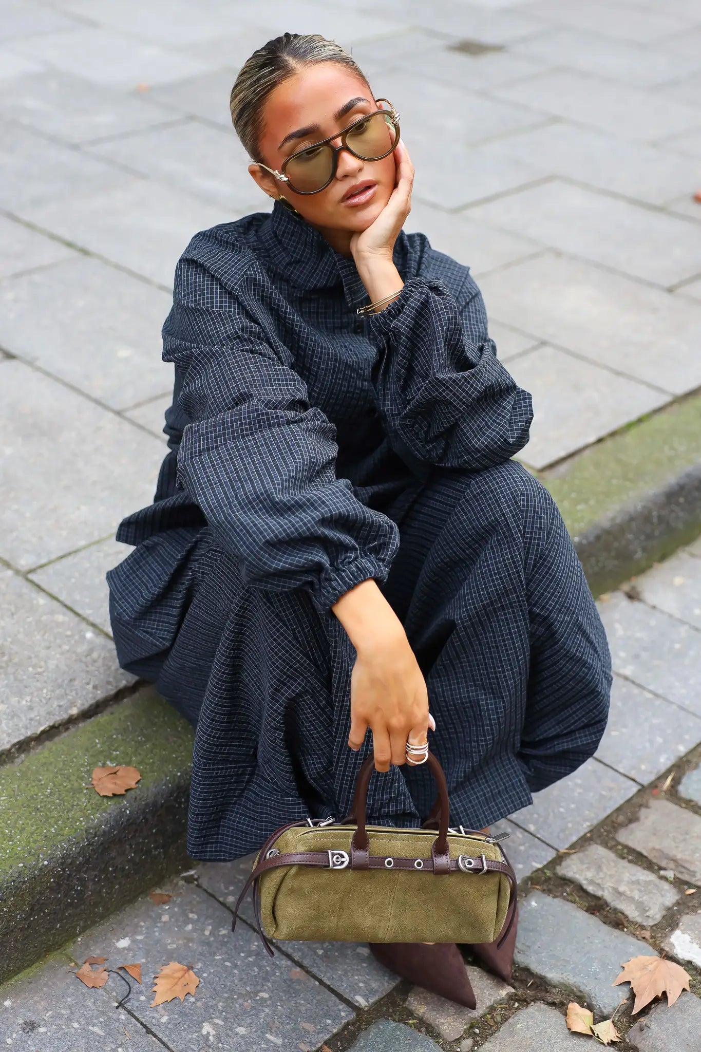 Khaki suède tas met bruine leren details gedragen bij een donkerblauwe oversized outfit terwijl het model op een trap zit