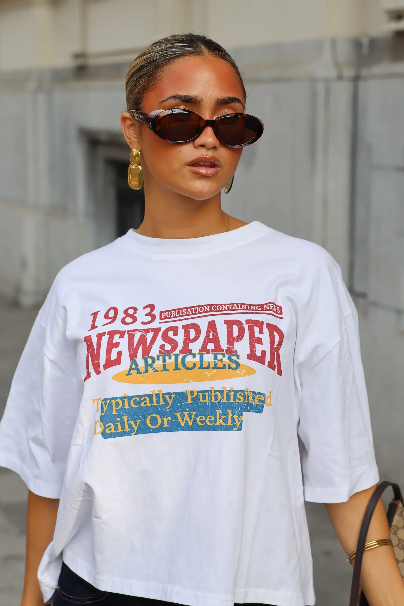Wit T-shirt met 1983 Newspaper Articles print in rood geel en blauw gedragen met een zwarte jeans en bruine zonnebril