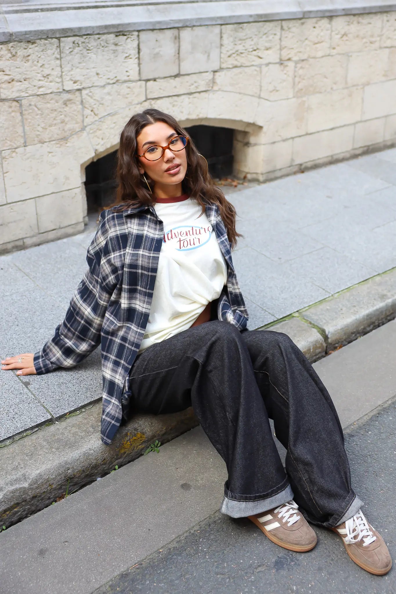 Model zit op de stoep in een crème Adventure longsleeve met donkerblauwe geruite blouse en zwarte wijde jeans.