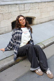 Model zit op de stoep in een crème Adventure longsleeve met donkerblauwe geruite blouse en zwarte wijde jeans.