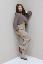 Model leunt tegen muur in taupe pantalon en grijze trui