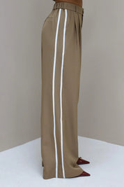 Model poseert in hoek met taupe pantalon met witte bies en hakken