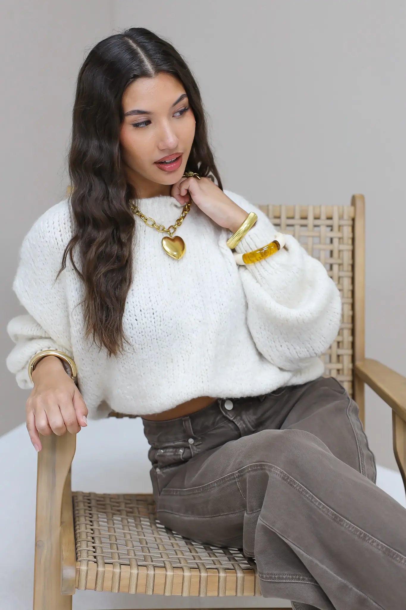 Model zit op houten stoel in witte fluffy trui met bruine broek