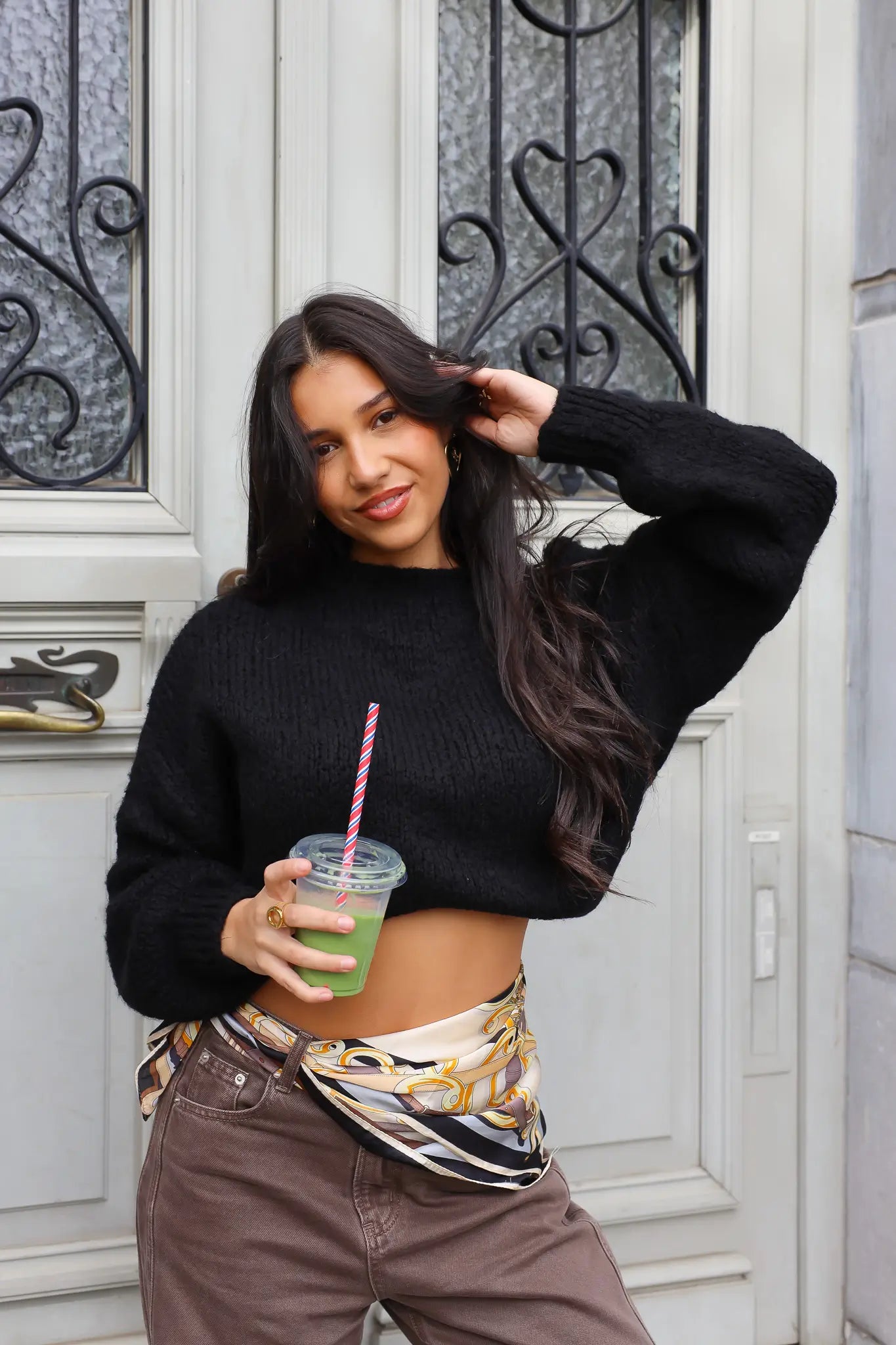 Model draagt zwarte cropped trui met sjaalriem en bruine broek buiten bij deur