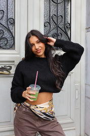 Model draagt zwarte cropped trui met sjaalriem en bruine broek buiten bij deur