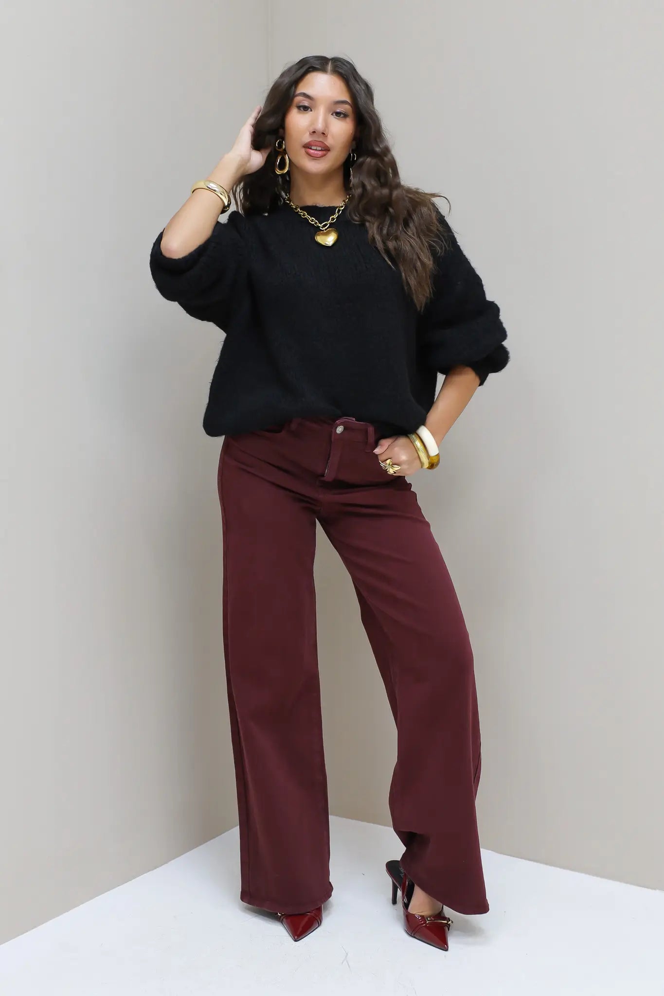 Model leunt tegen muur in zwarte cropped trui en bordeaux broek