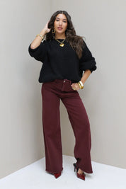 Model leunt tegen muur in zwarte cropped trui en bordeaux broek