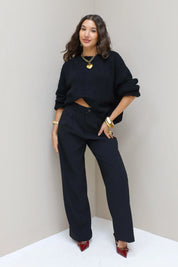 Model draagt zwarte pantalon met zwarte trui en hakken